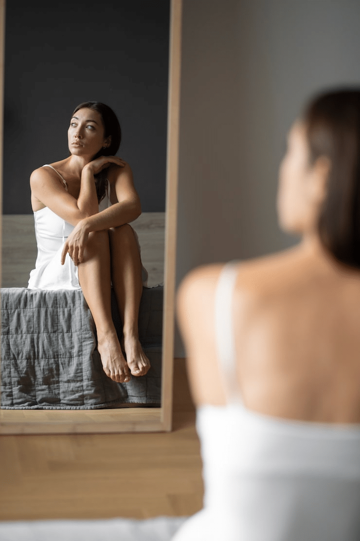 A young woman with low self-esteem looking away from a mirror.