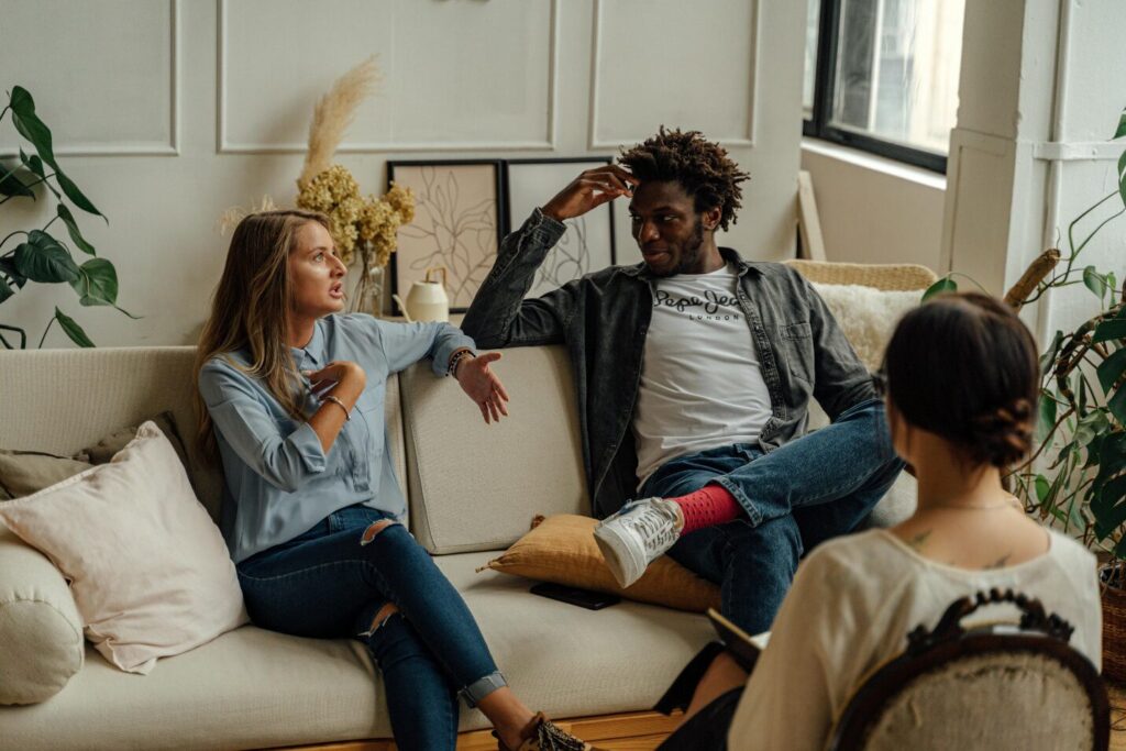 A couple sits on a couch in a room full of plants and art while another person sits opposite on a chair holding a notebook, symbolizing the comfort that comes with finding the right therapist