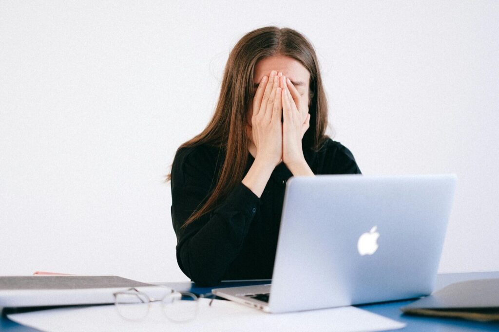 A woman holds her face in her hands as she sits at her desk with an open Macbook in front of her with scattered paperwork, files, and spectacles before starting online therapy