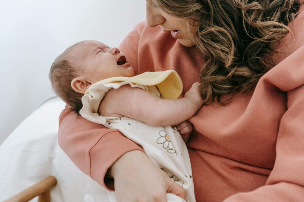 A woman in a pink sweatshirt holds a crying infant close to her body while looking down, showing how postpartum depression can sometimes go unrecognised