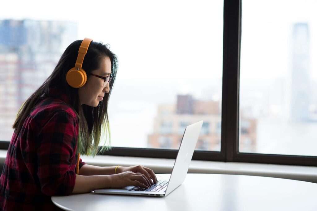 A woman in a red and black chequered shirt and yellow headphones sits by a large window while using a laptop on a round white table