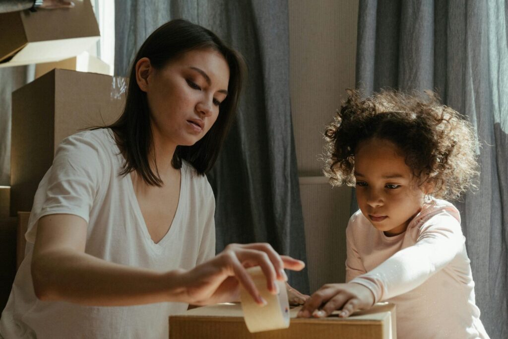 A woman in a white t-shirt and a young child in a light pink full-sleeved t-shirt pack a carton with tape while others are stacked behind them