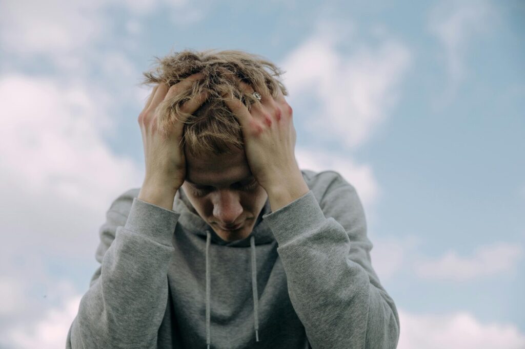 A young male adult sits with his head in his hands while wearing a grey hoodie with a cloudy blue sky above him