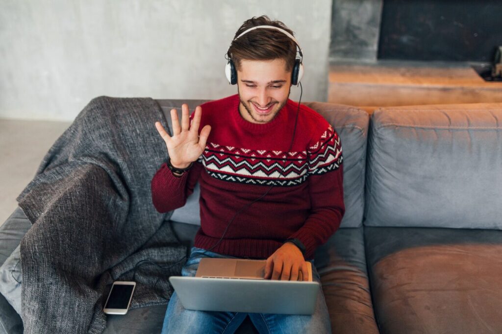 A man smiles as he waves to his therapist during an online session from his couch.