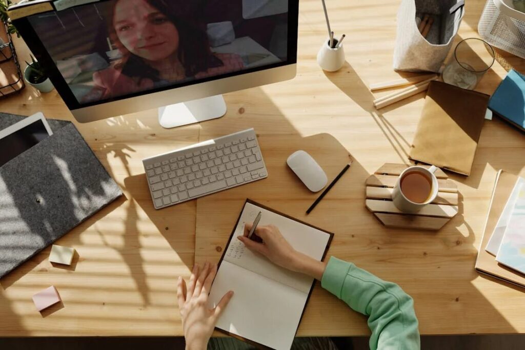 An image of a person writing something in a notebook while sitting in front of a computer