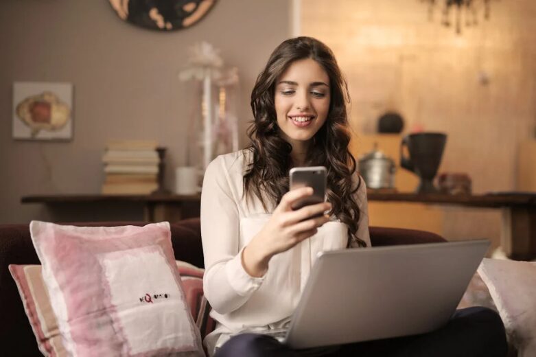 An image of a woman looking at her phone while sitting with a laptop on her lap