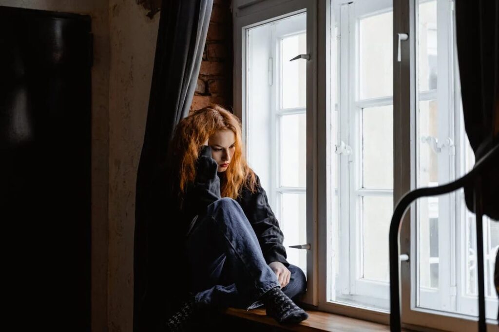 An image of a woman sitting by a windowsill holding her hair, looking sad